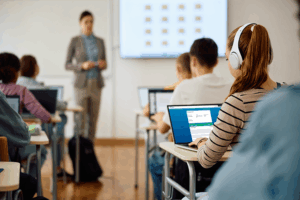 A student is in class wearing headphones and using a laptop with CLEAR Connect on it while a teacher is giving a lesson.