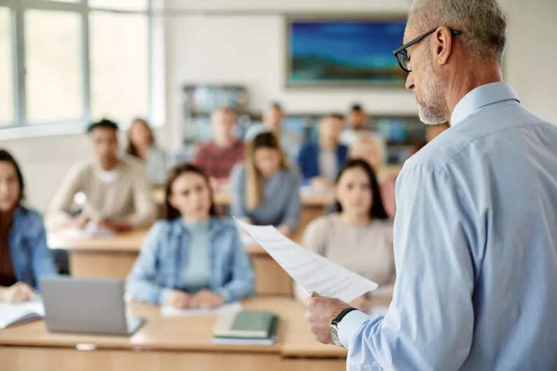 Male Teacher reads from paper while giving lecture to his students in the classroom.